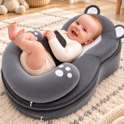 A baby lying on a plush gray baby support cushion with bear ears, placed on a textured cream rug in a bright indoor room.